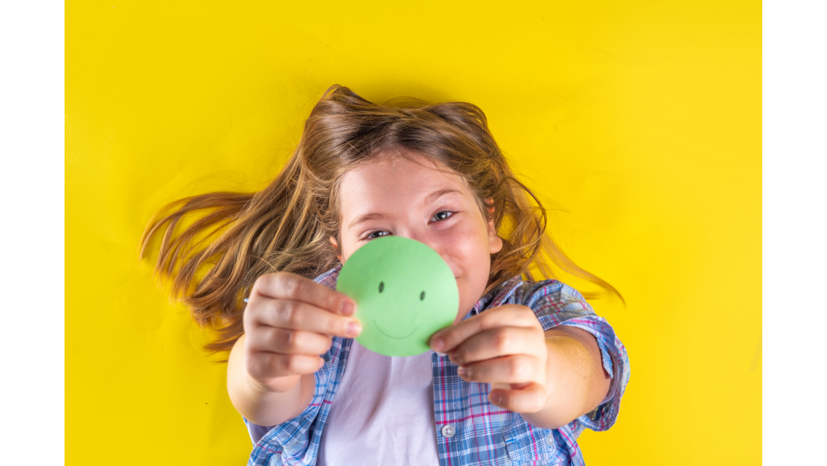 A young girl with long brown hair smiles against a yellow background while holding a green smiley face cutout piece of paper.