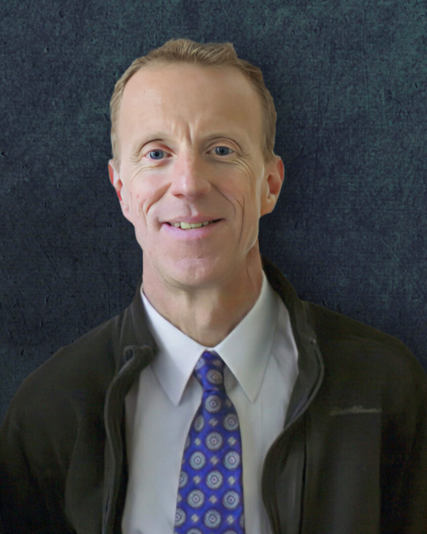 Closeup of a smiling middle aged white man with short-cropped light brown hair, wearing a white shirt, dark blue tie, and fleece jacket 