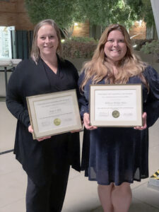 Two blonde women stand smiling and holding framed diplomas. The woman on the left is in a black top, sweater, and slacks, and the woman on the right is in a navy blue knee-length dress