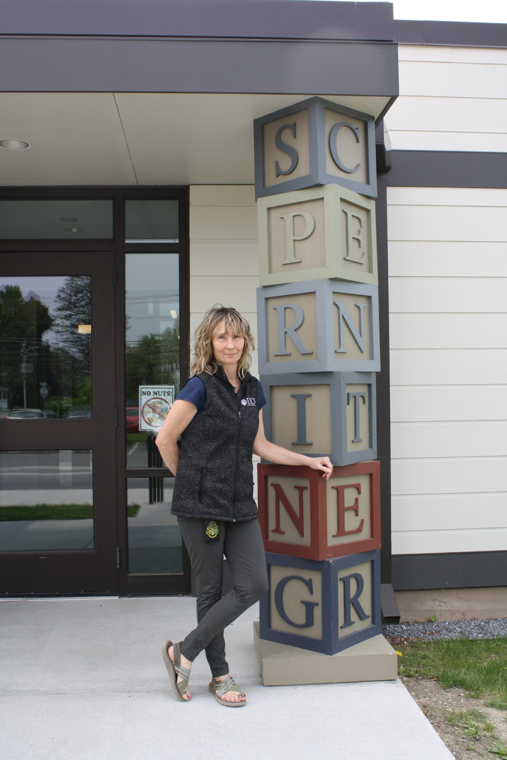 A thin woman with curly blonde hair wearing a black vest and grey pants stands in front of a modern building with a pillar spelling "spring" that looks like children's building blocks.