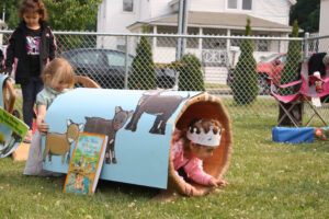 A group of preschool children and toddlers play in a fenced in yard, climbing through a kid-sized fabric tube with hand-painted pictures of animals on it and a picture book propped up against it.