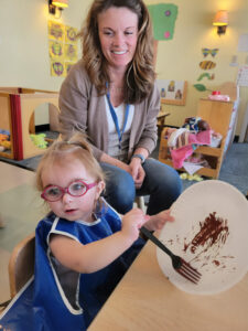 A closeup of a blonde toddler in a blue smock and red glasses sitting at a kid-size table and holding up a fork and paper plate on which she has “painted” with what appears to be chocolate Seated on a stool next to her is a smiling young blonde woman with a beige cardigan, white t-shirt, and blue jeans