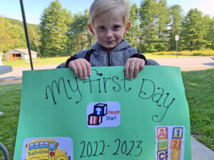 A closeup of a preschool age blond boy holding up a green sign that says “My first day of school” and the Head Start logo of red and blue stacked blocks. He is standing on the grass with trees and mountains in the background