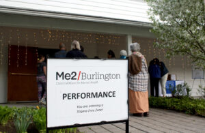 Photo of sign in front of a line of people, sign reads "Me2/Burlington Perfomance