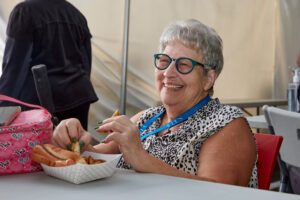 A smiling white woman with short gray hair, blue-framed glasses, and a black and white patterned shirt sits at a table with a tray of French fries in front of her.