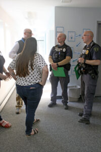 a heavyset young white woman with long brown hair wearing a white t-shirt with black polka dots and blue jeans, is seen with her back to the camera. Before her are three men; at left, a white man with a shaved head and eyeglasses, wearing a blue fleece vest and white dress shirt, who is in conversation with the two uniformed police officers, both white men with short cropped hair, standing opposite him and the woman.