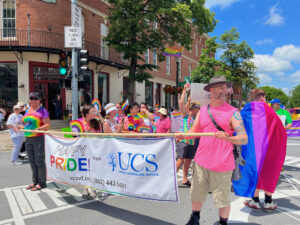 A group of people, many wearing rainbow tie-dyed tshirts or wrapped in rainbow flags, walk behind a man in a pink sleeveless shirt and khaki shorts and a woman in a rainbow tie-dyed t-shirt who are are carrying a long banner between on a pole. The banner reads “Happy Pride” and displays the UCS logo.