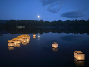 A deep blue nighttime scene of a lake with trees across the far end and the moon glowing above. On the lake are floating numerous cube-shaped paper lanterns, glowing with candlelight.