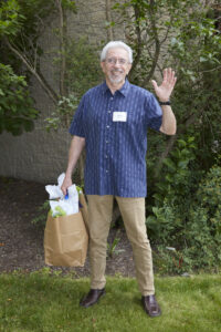 Older man with gray cropped hair standing and waving at the camera with a big bag in hand