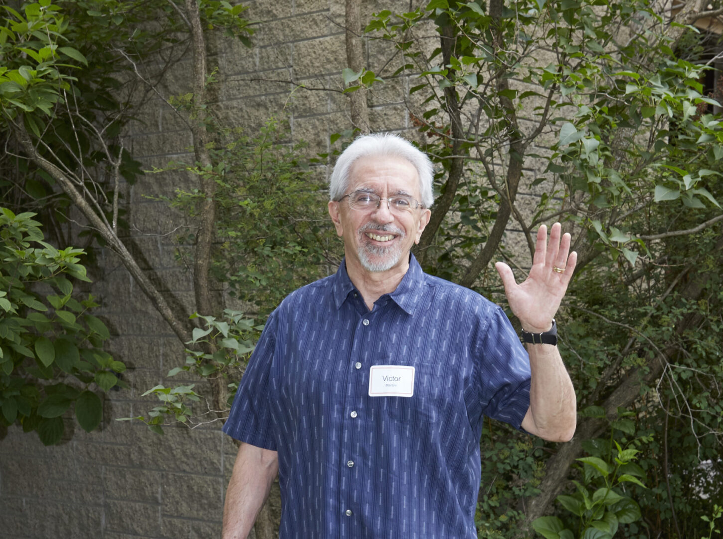 Older man with gray cropped hair standing and waving at the camera with a big bag in hand