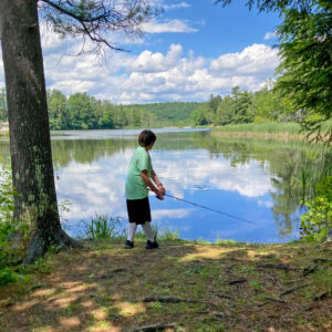 a young white boy in a mint green t-shirt, dark blue shorts, white knee-socks and sneakers is seen standing under a tree at the edge of a lake with his back to camera. He is holding a fishing rod and appears to be preparing to cast his line.
