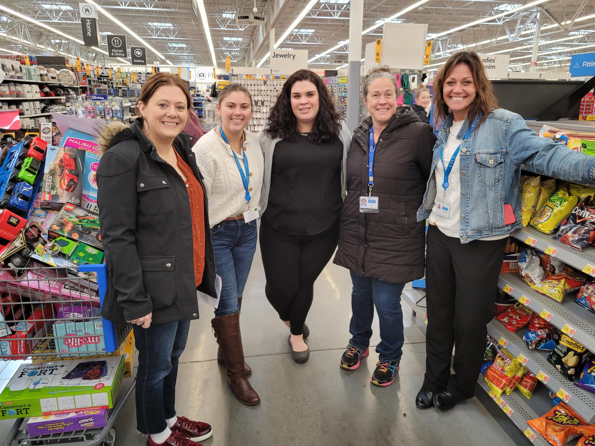 Five smiling women, some wearing blue name tag lanyards, stand together in a brightly lit store setting. They are surrounded by brightly colored store items. Left to right: Woman with orange sweater and red hair, woman with white shirt with light brown hair tied back, woman with black curly hair wearing all black, woman with silver-brown curly hair wearing a brown winter coat, and a woman with brown hair wearing a white shirt and denim jacket.