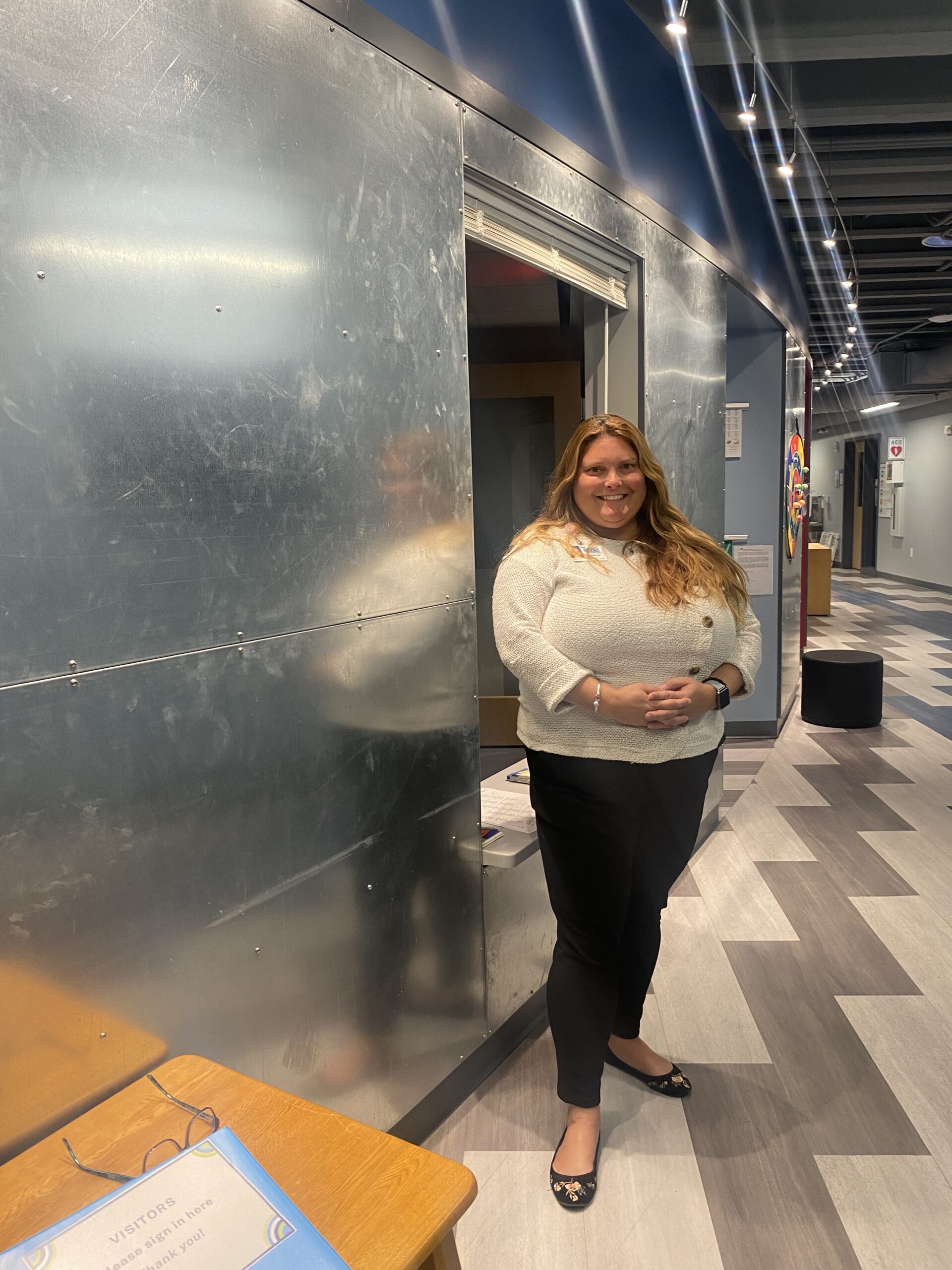 A woman with long red hair wearing a white sweater, black pants, and black flats with a floral pattern smiles and stands next to a modern metal wall in a hallway with alternating gray and white floor tiles.