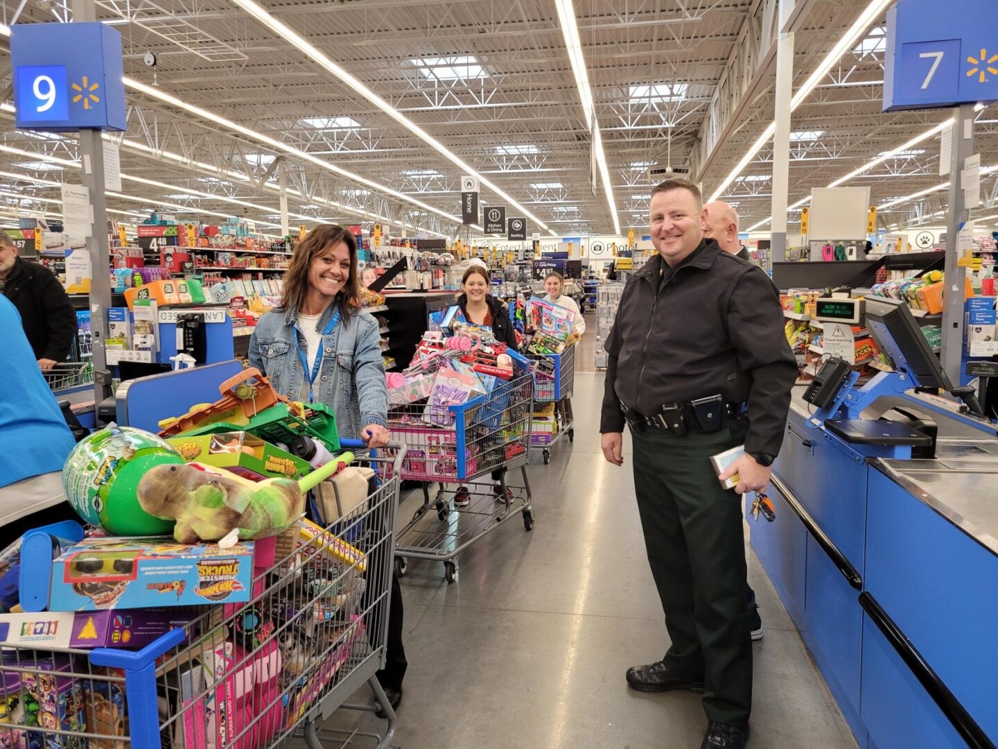 Three women lined up with shopping carts filled with bright colored toys and children's items smile next to a smiling police officer wearing black. The group is in a brightly lit big store with checkout lane markers numbered 7 and 9. The markers are blue with white numbers and have a yellow star walmart logo. 