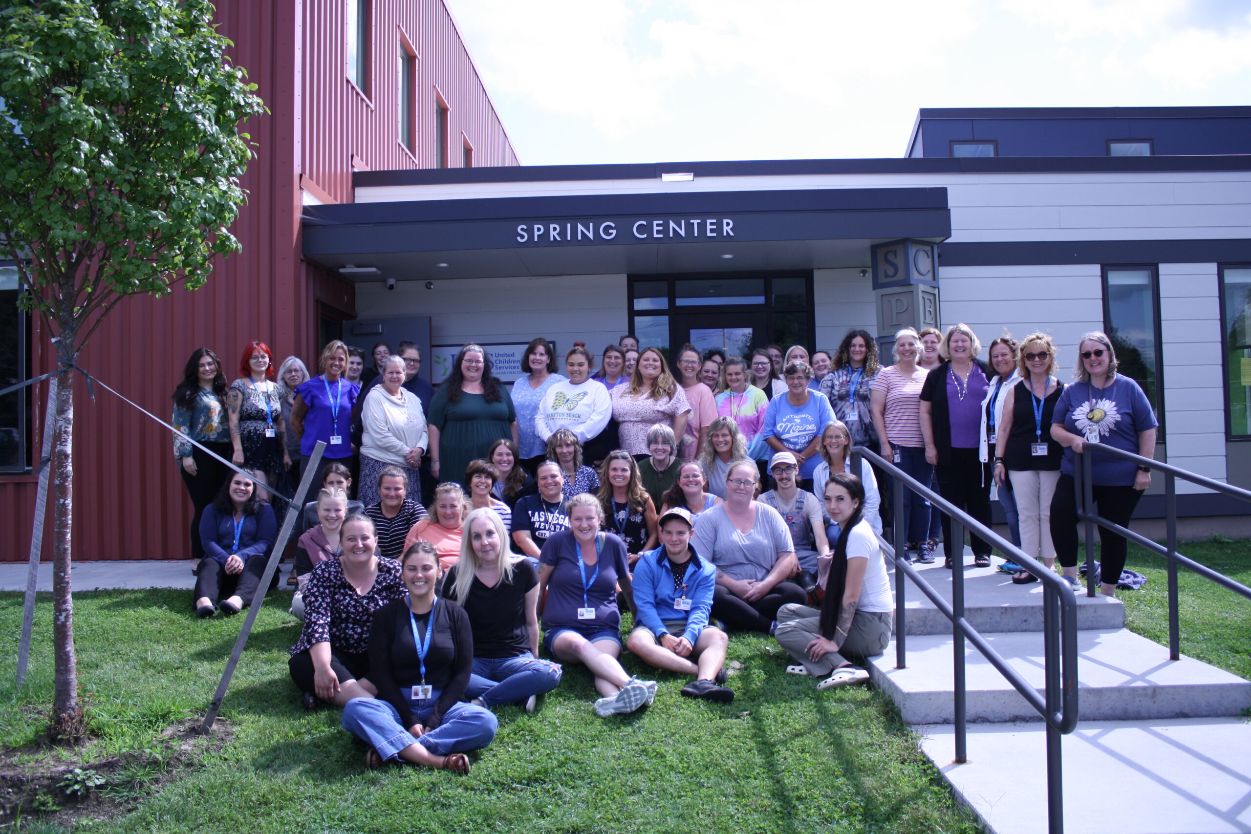 large group of adults smiles in front of a modern building with Spring Center on the awning. Some people are sitting on the grass and those in the back of the photo are standing.