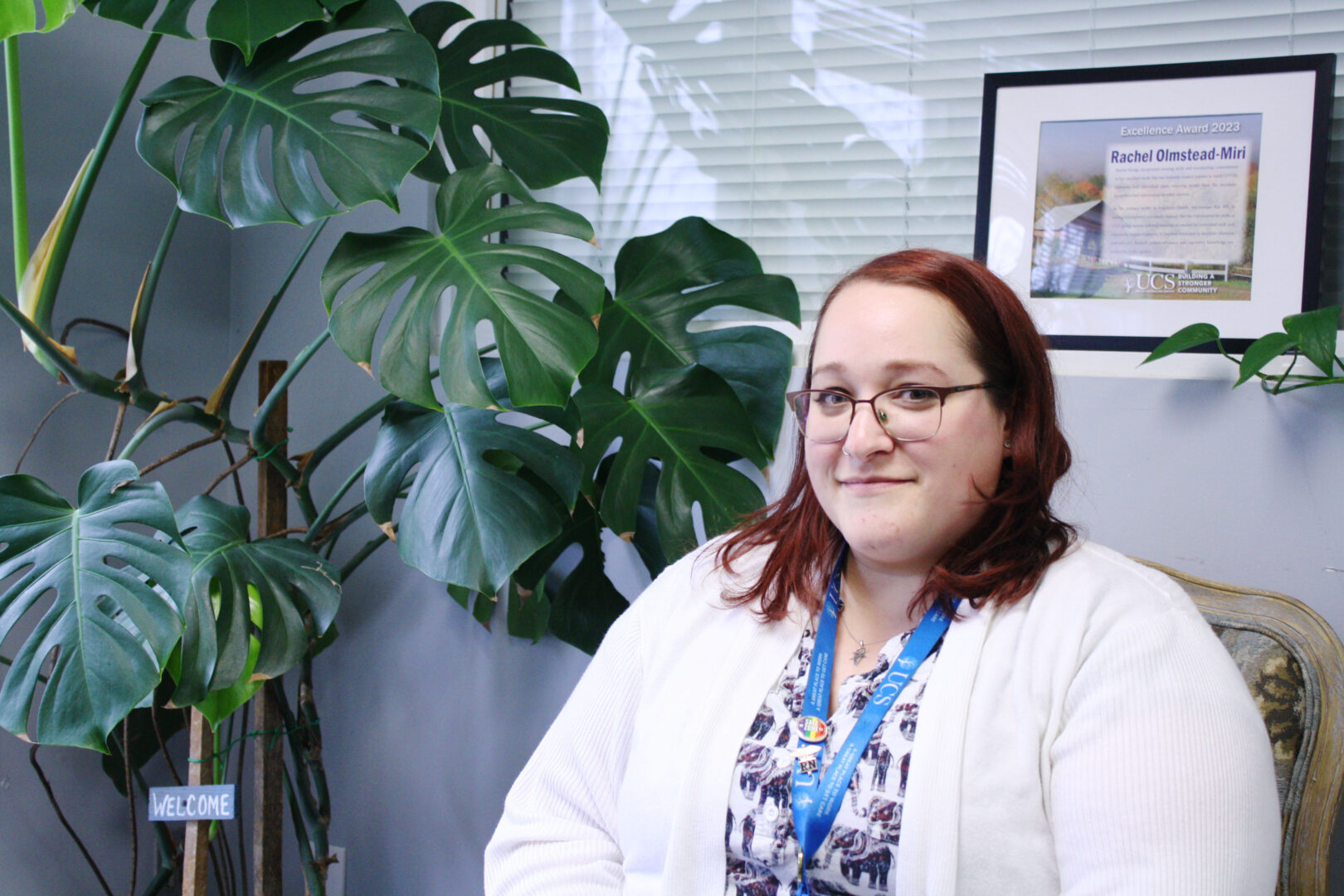 A woman with medium length red hair and glasses wearing a white sweater and floral pattern shirt sits with a monstera plant behind her.