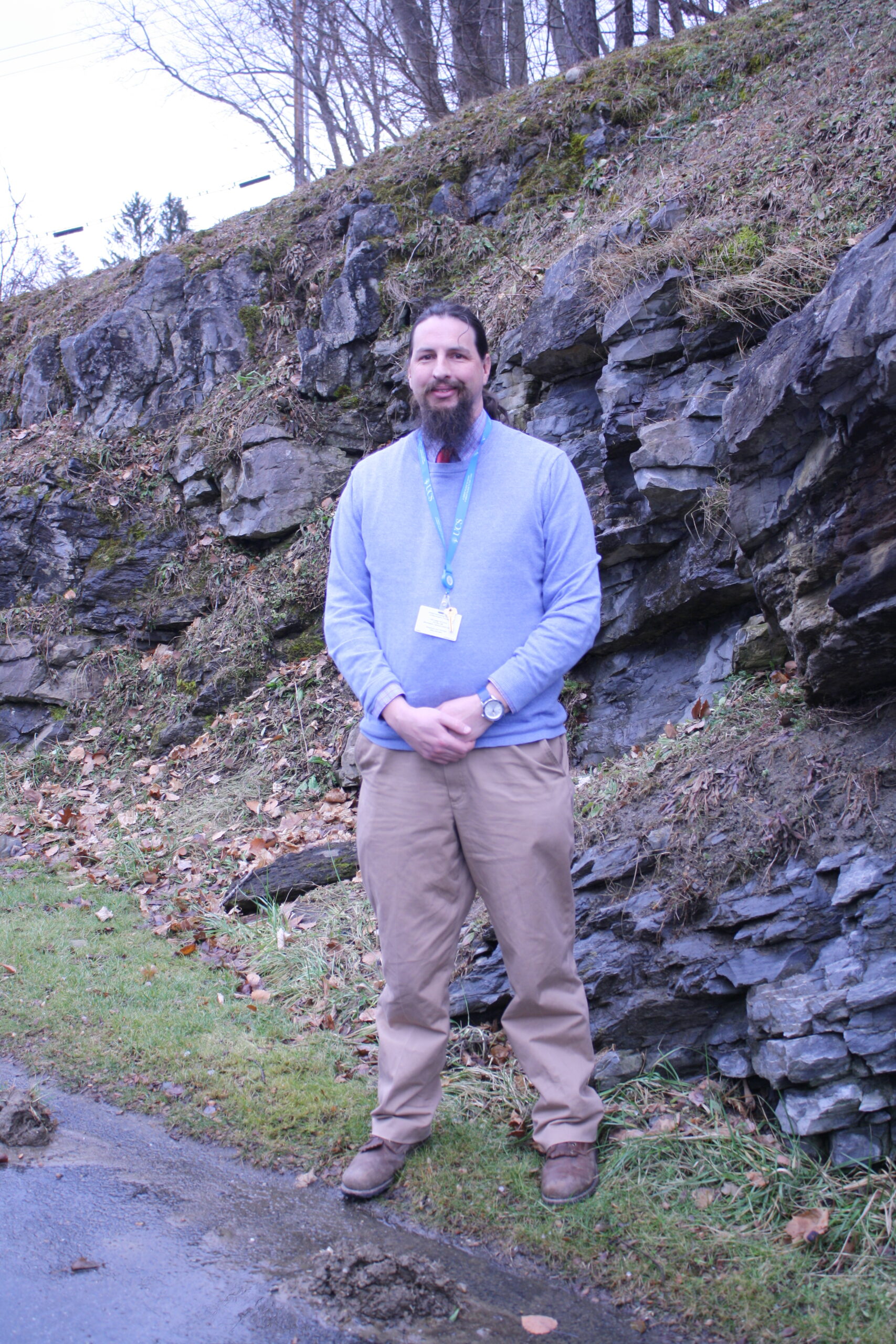 A man with long brown hair and a beard wearing a light blue sweater and tan pants stands in front of a rocky ledge.