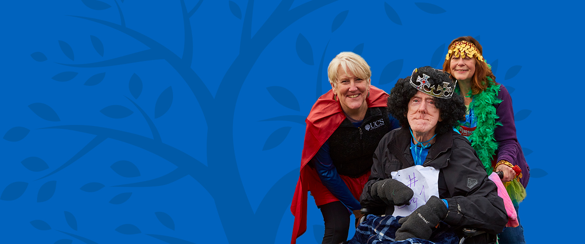 One older man in a wheel chair in a curly wig next to two women smiling in costume