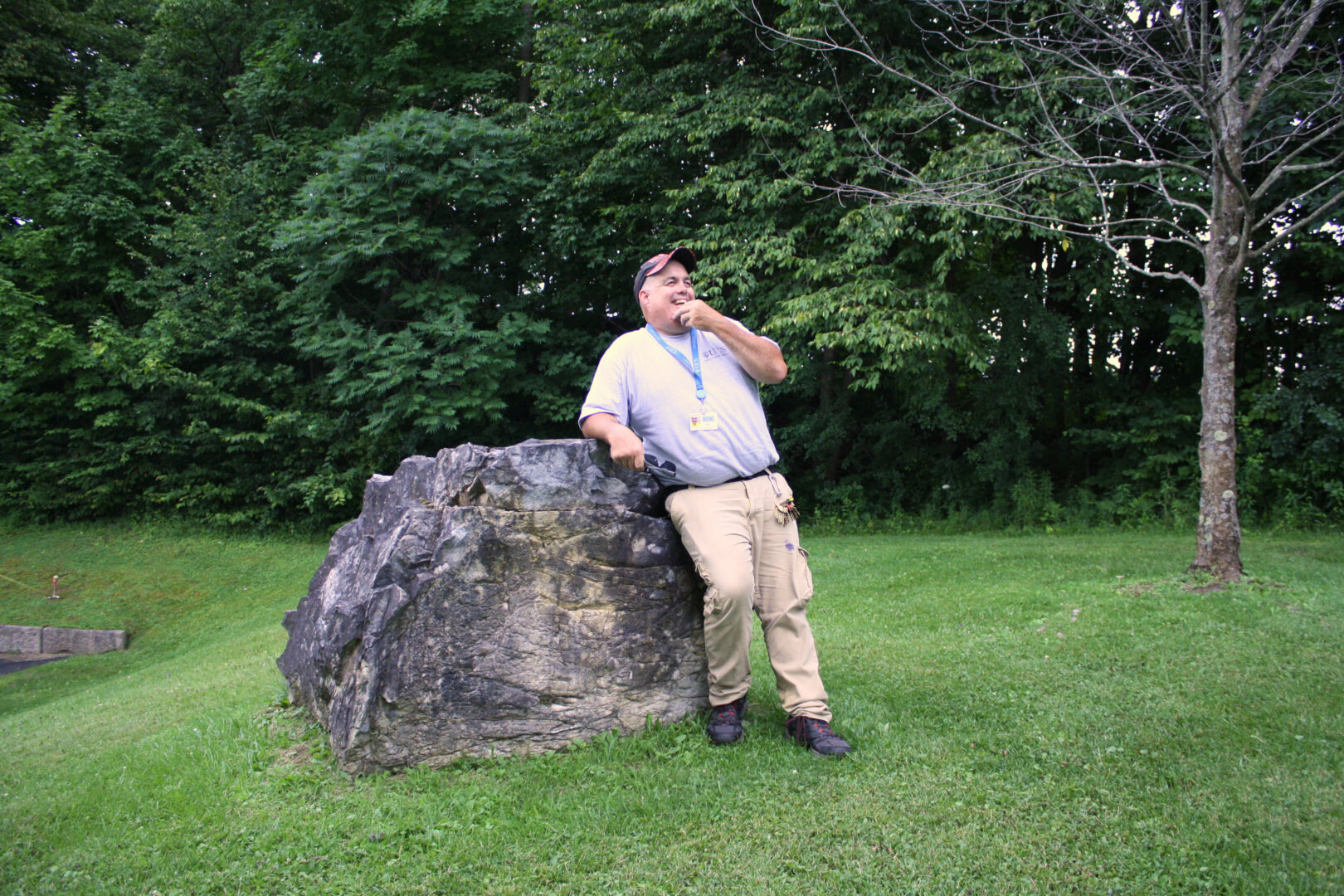 A smiling man with his left hand up to his chin leans against a boulder. Background has green grass and trees, and a small maple tree.