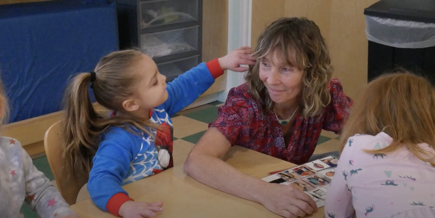 A little girl with light brown hair touches the hair of a woman with blonde hair who is smiling. Both are sitting at a desk.