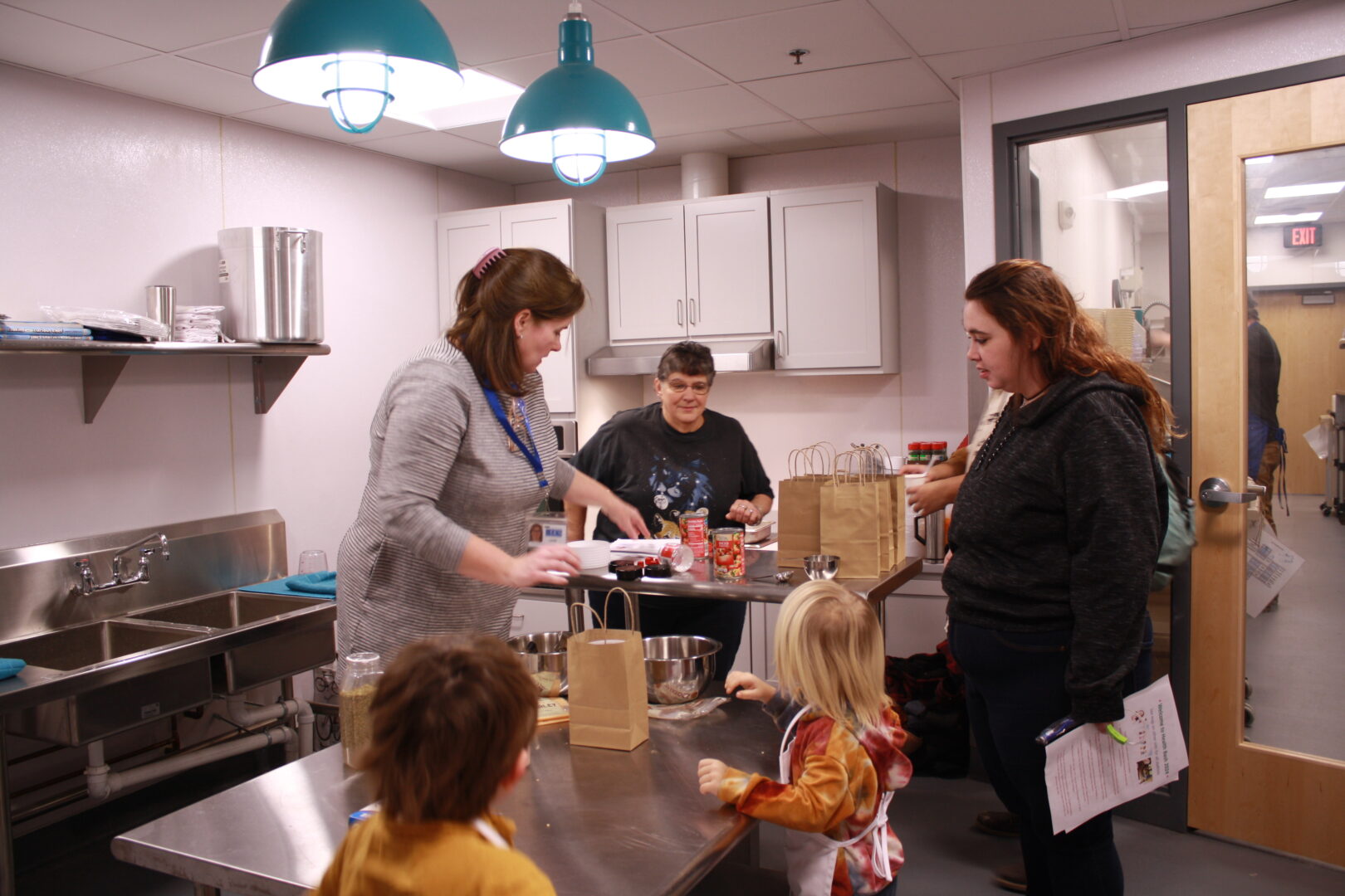 Two children work on a cooking project in a commercial style kitchen with three adults. 