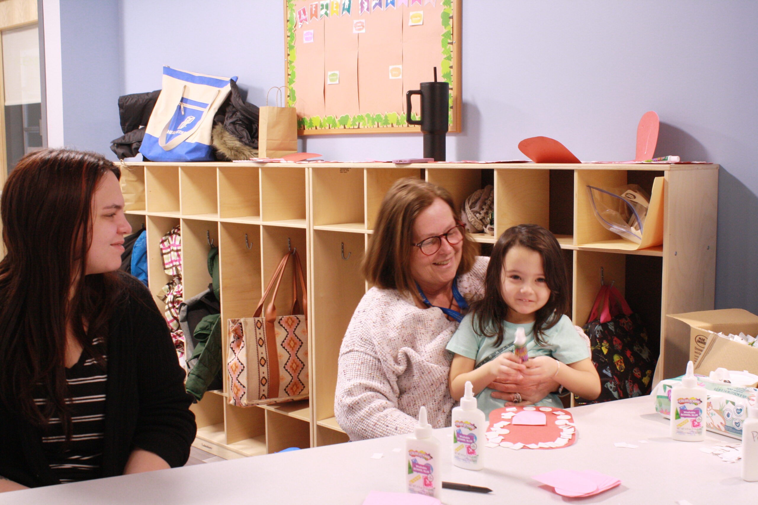 A woman smiles while holding a young girl on her lap. Another woman smiles and looks on. There is a classroom style room in the background with light colored wood cubbies.