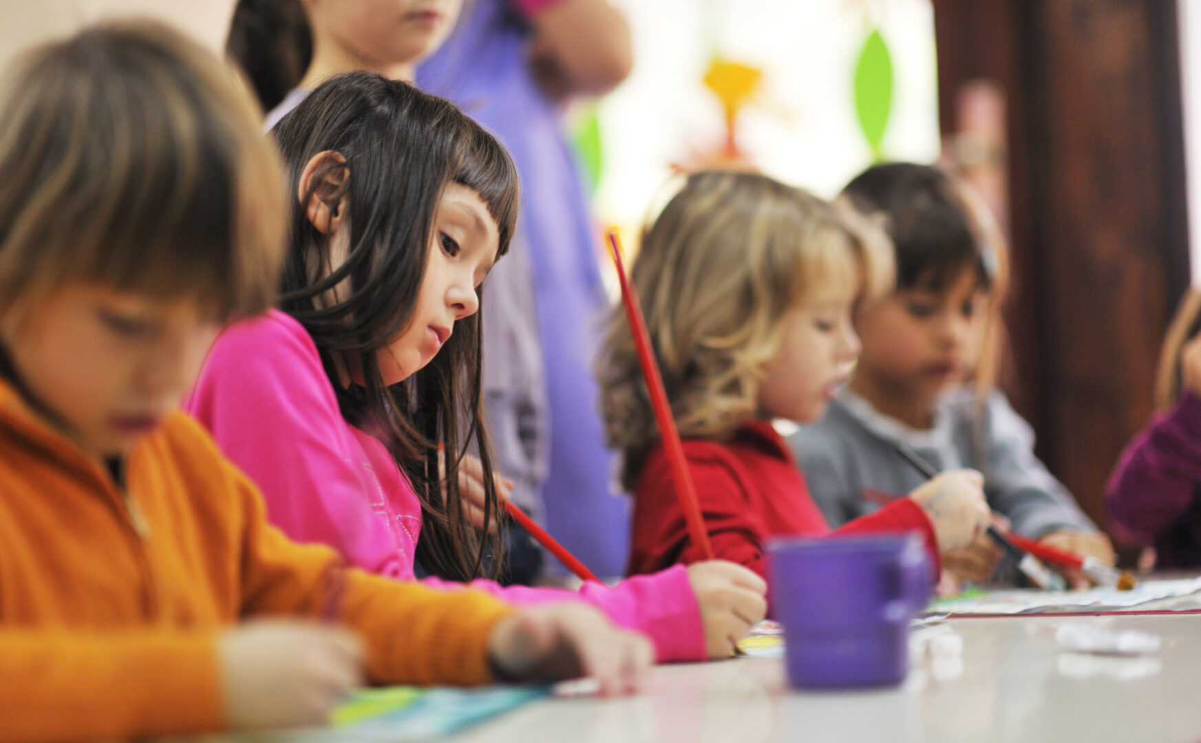 Three preschool age children creat art at a white table. From left ro right: young girl with long dark hair and bangs, young child with wavy blonde hair, and a young boy with short dark hair
