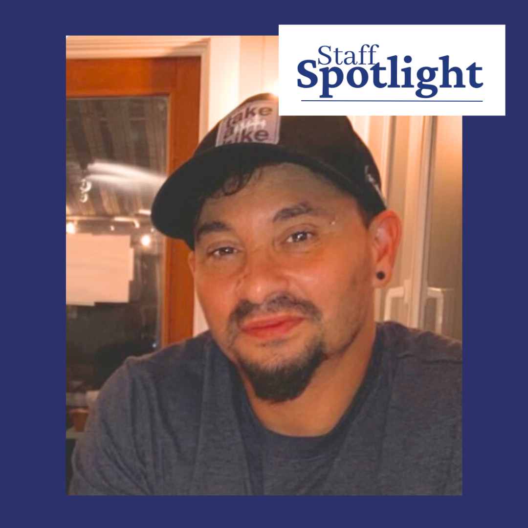 A man with a mustache and short beard wearing a black baseball cap. The image has a dark blue frame and there is a Staff Spotlight logo on the top right