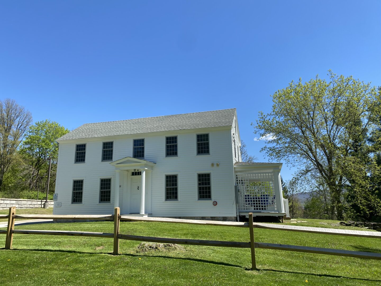 historic white house with a front door including two  pillars. Wooden fence and green grass in the foreground. Blue sky day.