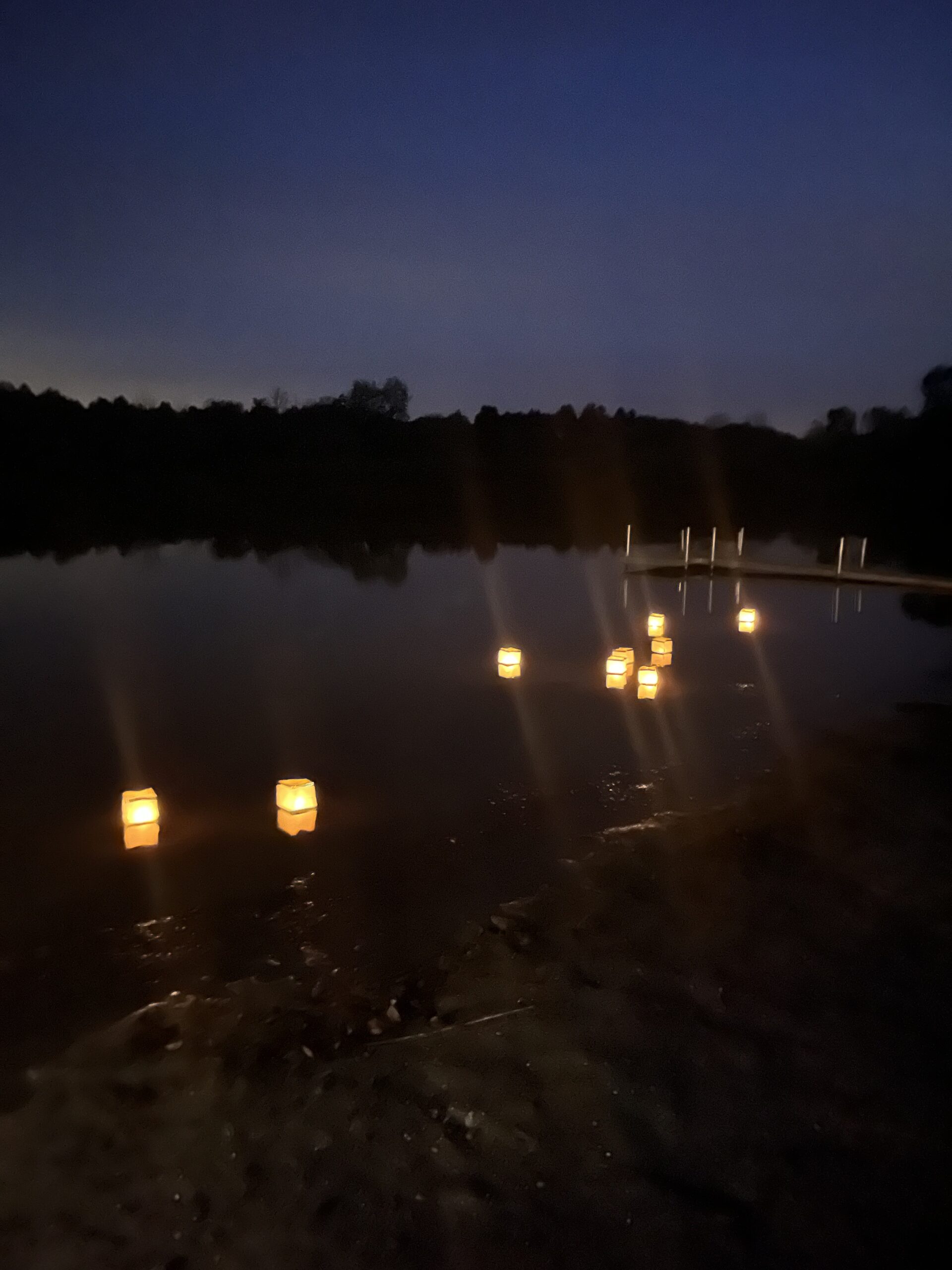 candlelit paper lanterns float on a dark late with a treeline and dark blue night sky in the background