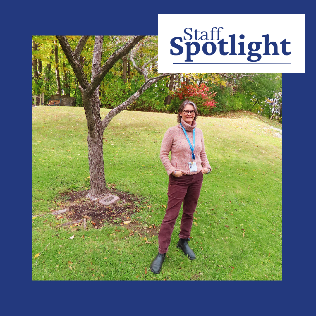 A woman with shoulder length silver hair wearing a pink turtleneck weater, ankle boots and maroon pants stands with her hands in her pockets next to a small tree. There is green grass as well. The top right has a logo image overlaid that says Staff Spotlight.