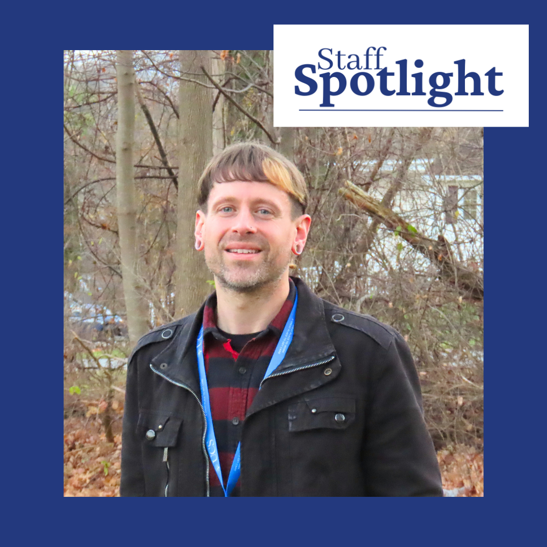 A person with shirt brown hair and a blonde streak wearing a black light jacket and a blue and red striped shirt and a blue lanyard smiles against a background of leafless trees. There is a frame that reads Staff spotlight in the right corner with a blue background.
