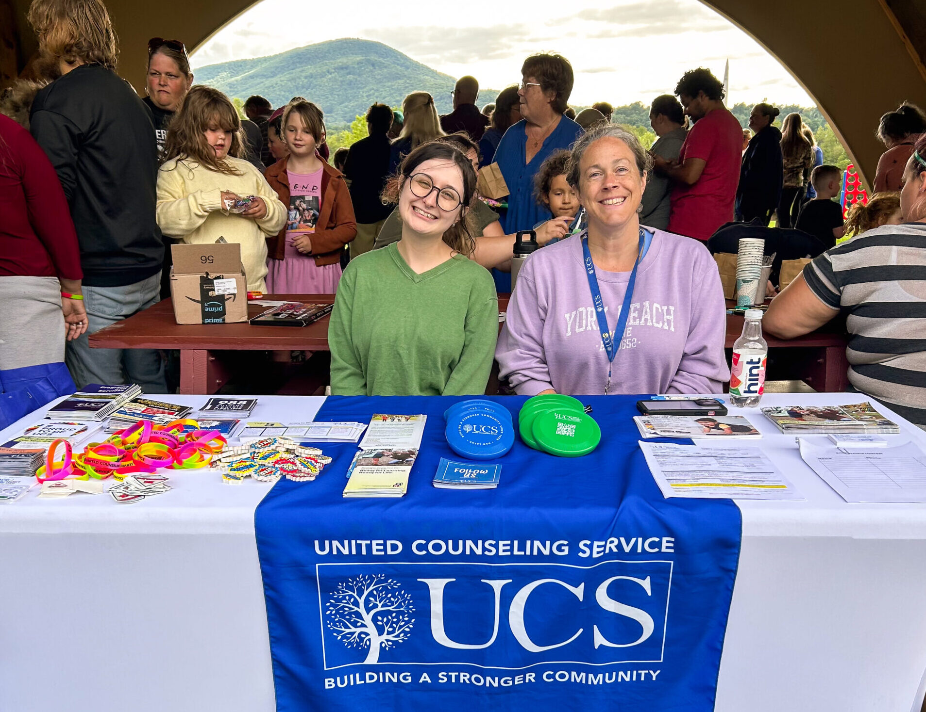 Two women sitting at a table at a public event with a blue banner with the text "UCS"