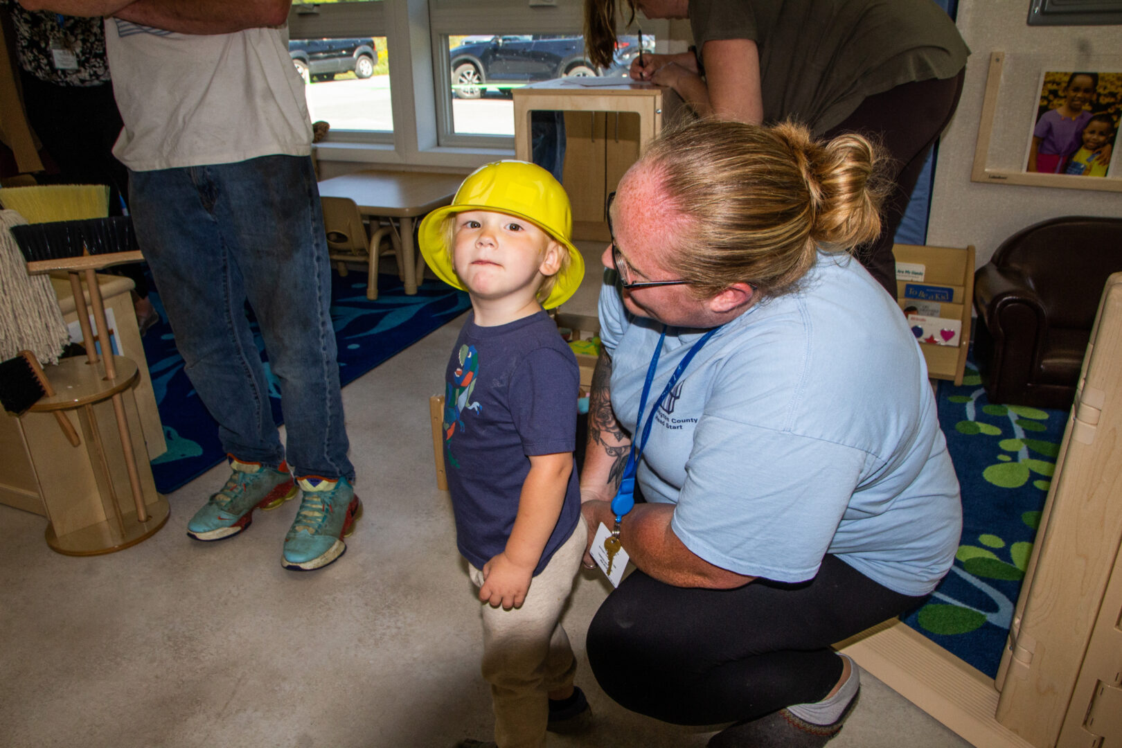 Toddler boy with yellow construction hat on looks at the camera while his mom kneels next to him looking at him