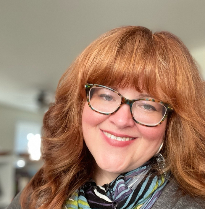 Women with long, red hair and glasses slightly tilts her head and smiles.