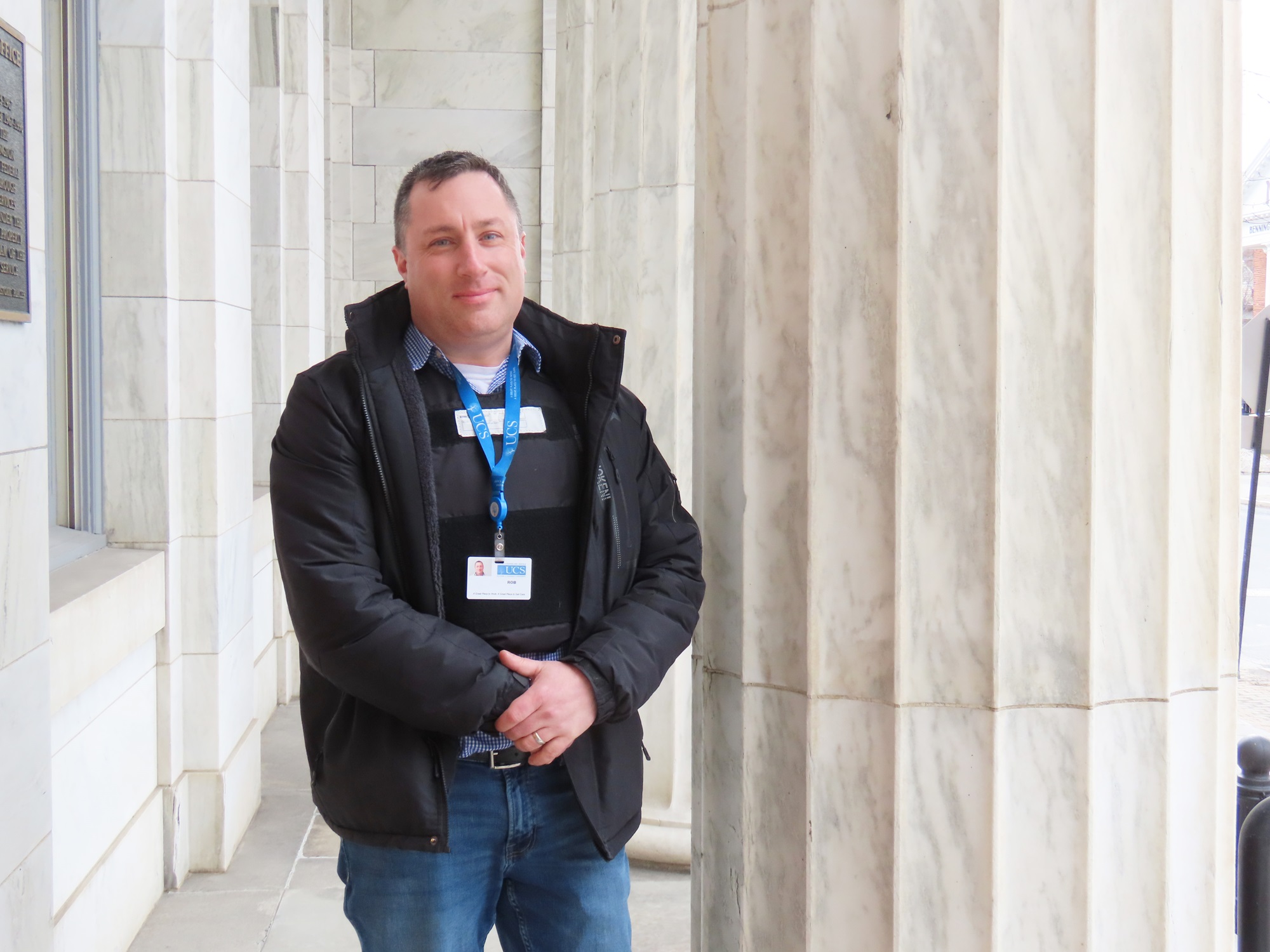 Man in police vest and black winter coat smiles in front of a marble pillar.