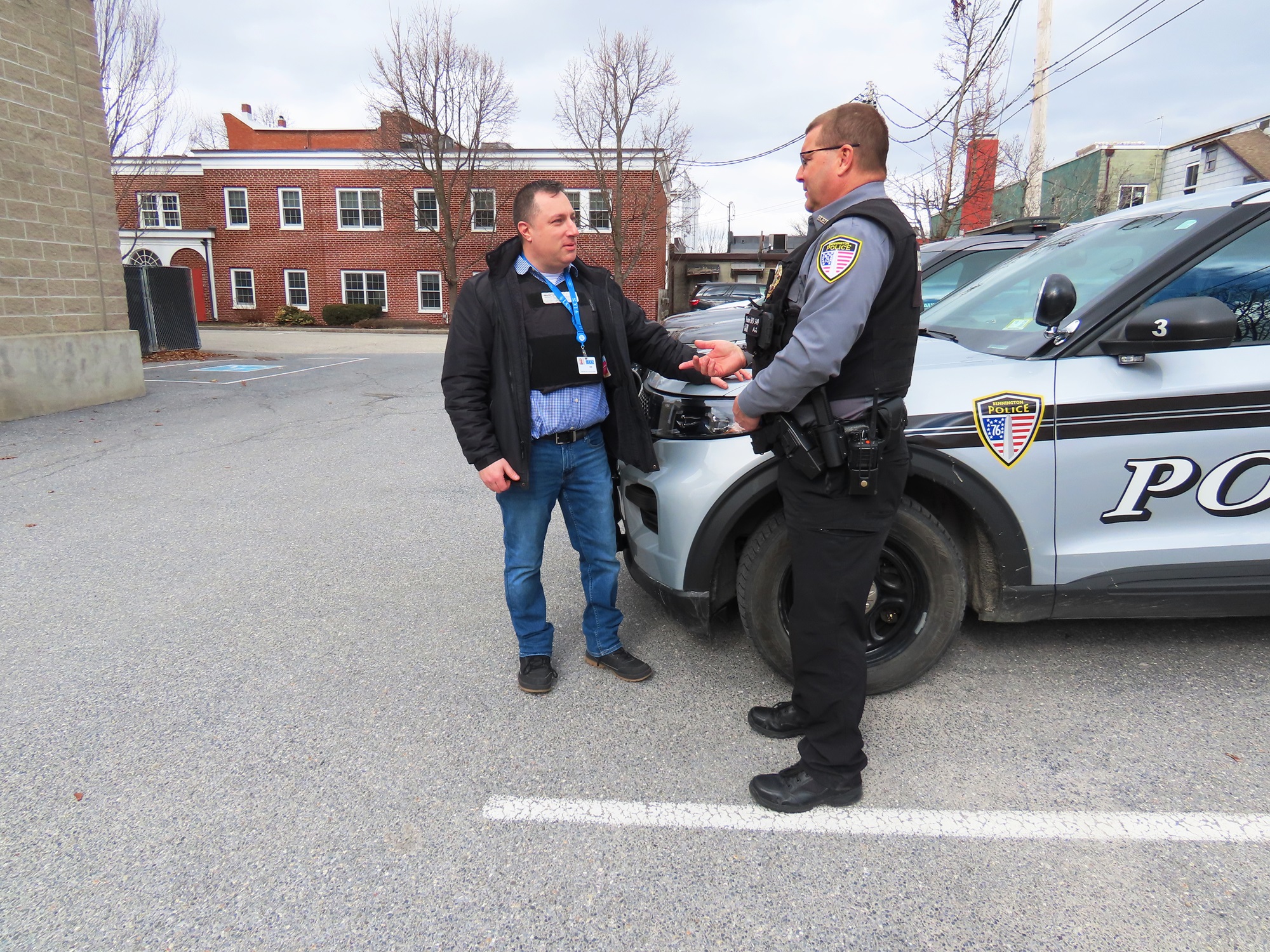 A law enforcement officer speaks with a man wearing a blue lanyard and police vest in front of a police cruiser.