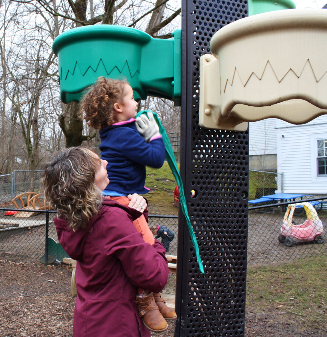 Woman wearing pink jacket holding up small child with blue jacket on a playground equipment