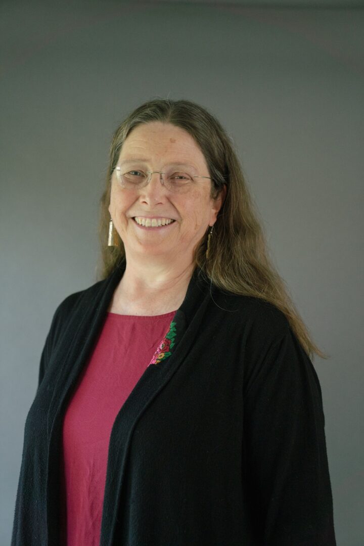 woman with long brunette hair wearing a pink/red shirt and black sweater and glasses smiles against a gray green background