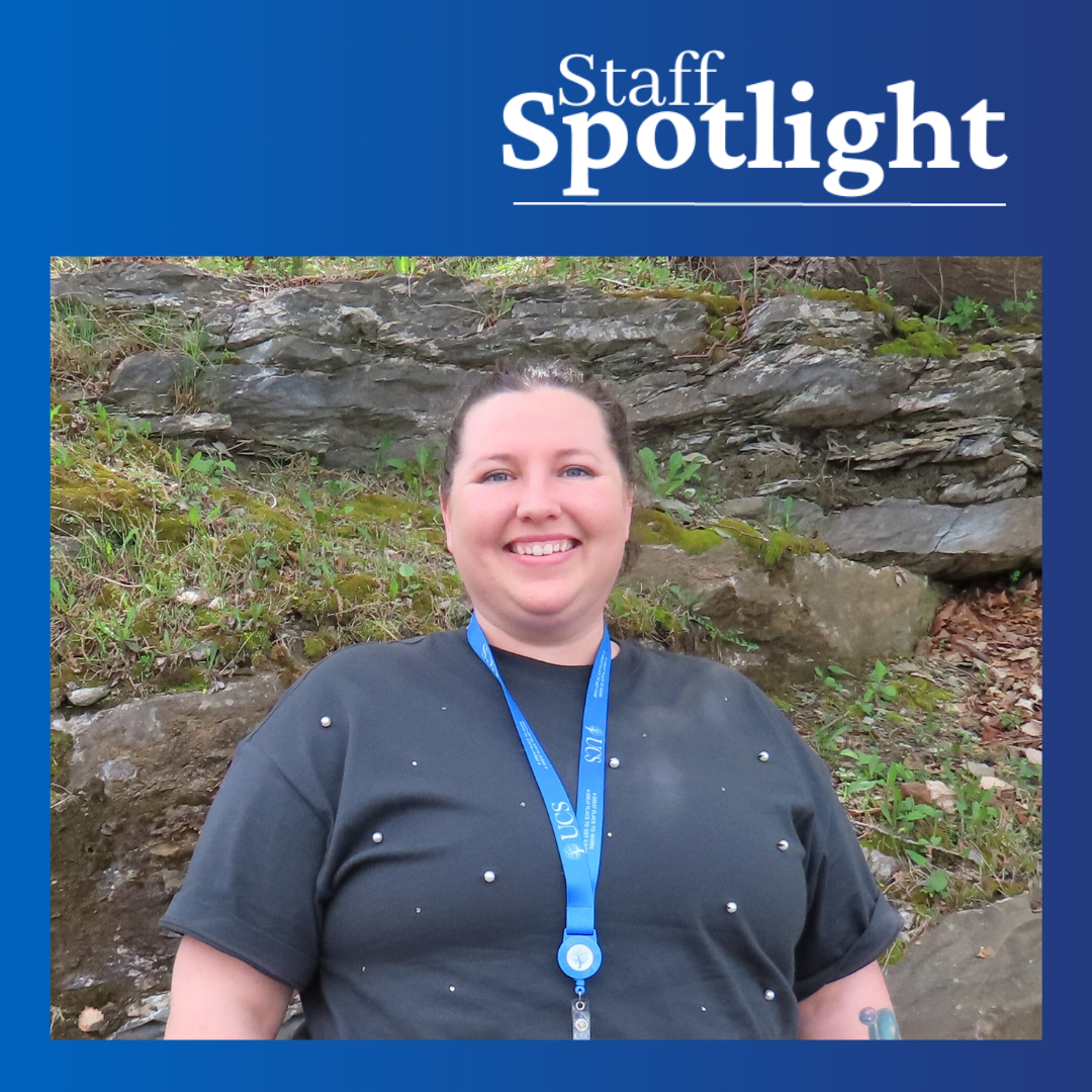 person wearing dark grey tee shirt with pearl decorations and wearing a blue lanyard while smiling in front of a rocky grassy background.