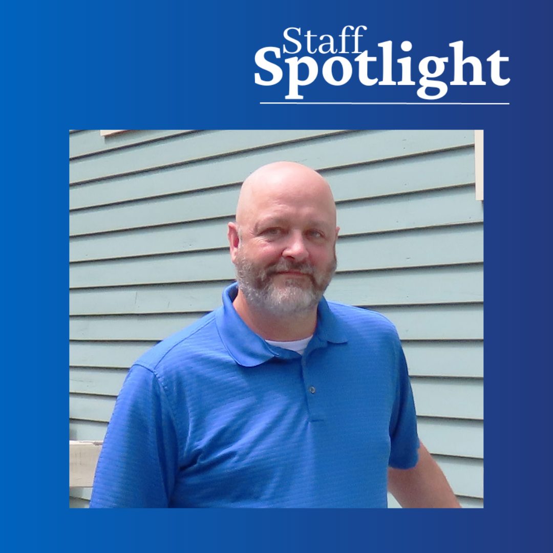Man with gray beard and mustache wearing blue polo shirt leanin on rail in front of a building with gray siding.