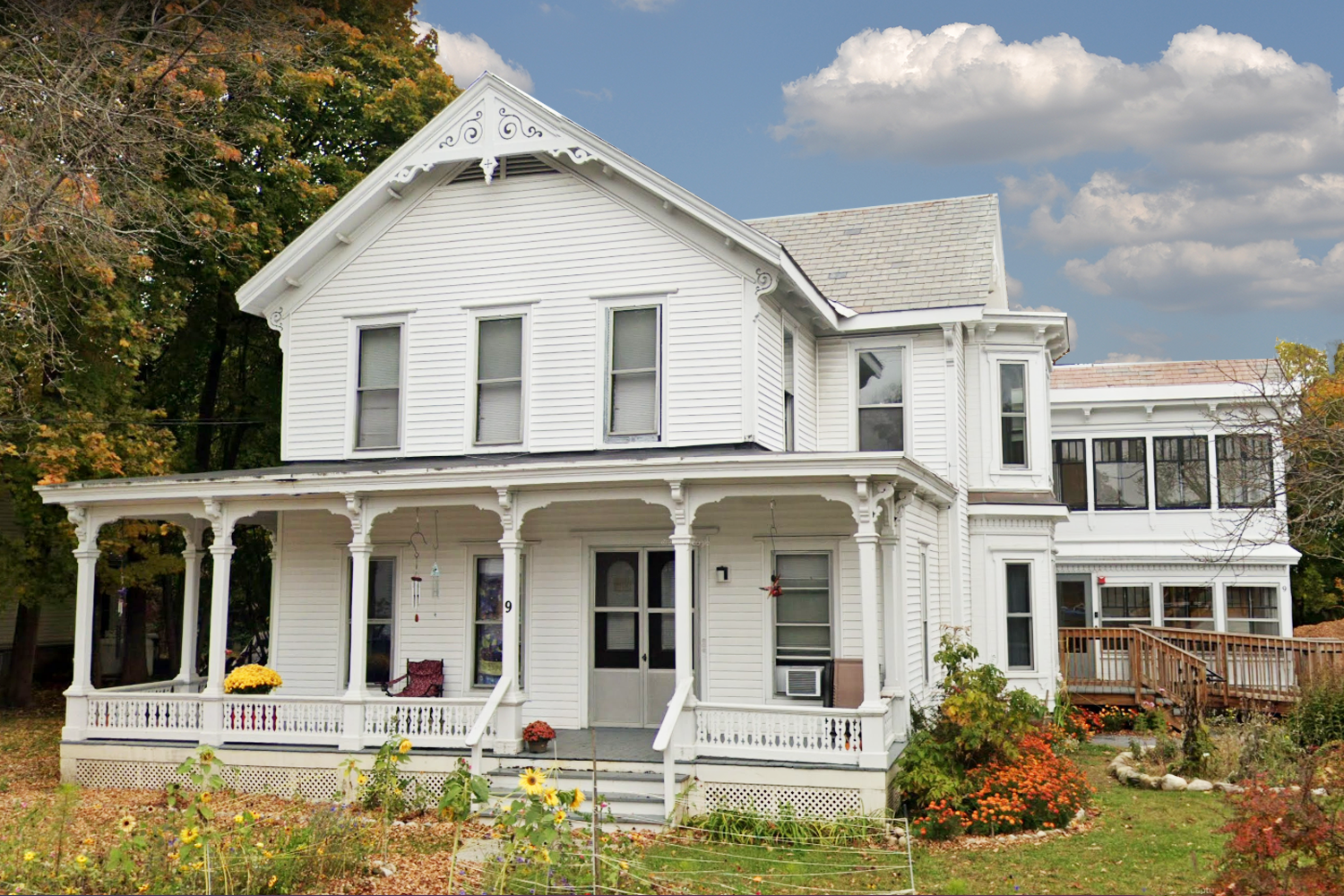 historic white house with six pillars and a front porch in an autumn setting with yellow mums on the porch and an orange bush on the side of the house. Sunny blue sky day with white clouds