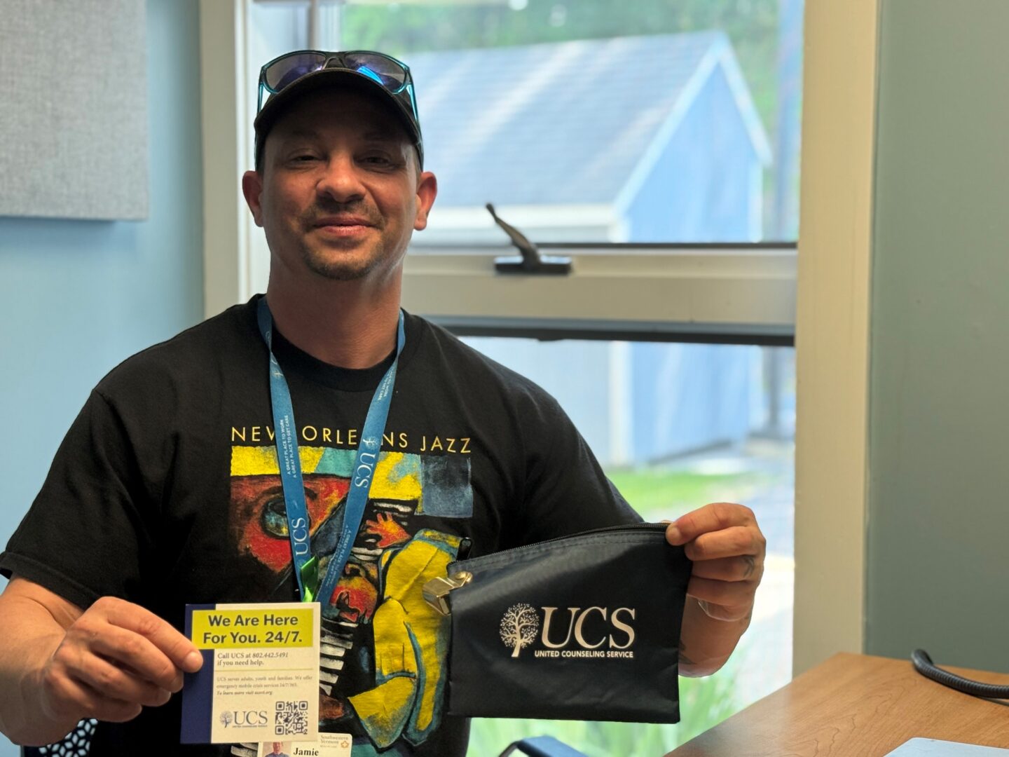 Smiling man wearing a baseball cap and sunglasses on his hat and a blue lanyard holds a blue bag with the UCS logo and a small blue, green, and white card.
