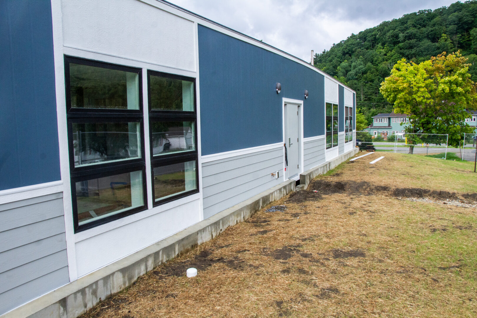 blue and white modular building with large windows sitting on a green-brown grassy area with green trees and a green hillside in the background