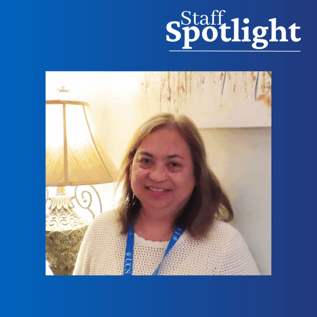 A smiling woman with long brown hair wearing a white shirt and blue lanyard stands in a gray room lit by an ornate lamp with a multicolored contemporary art piece in the background. Around the photo is a blue frame that says 'Staff Spotlight'.