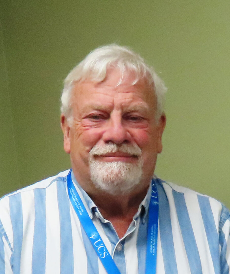 Man with white hair and white goatee wearing a blue and white striped shirt and blue lanyard in front of a green wall
