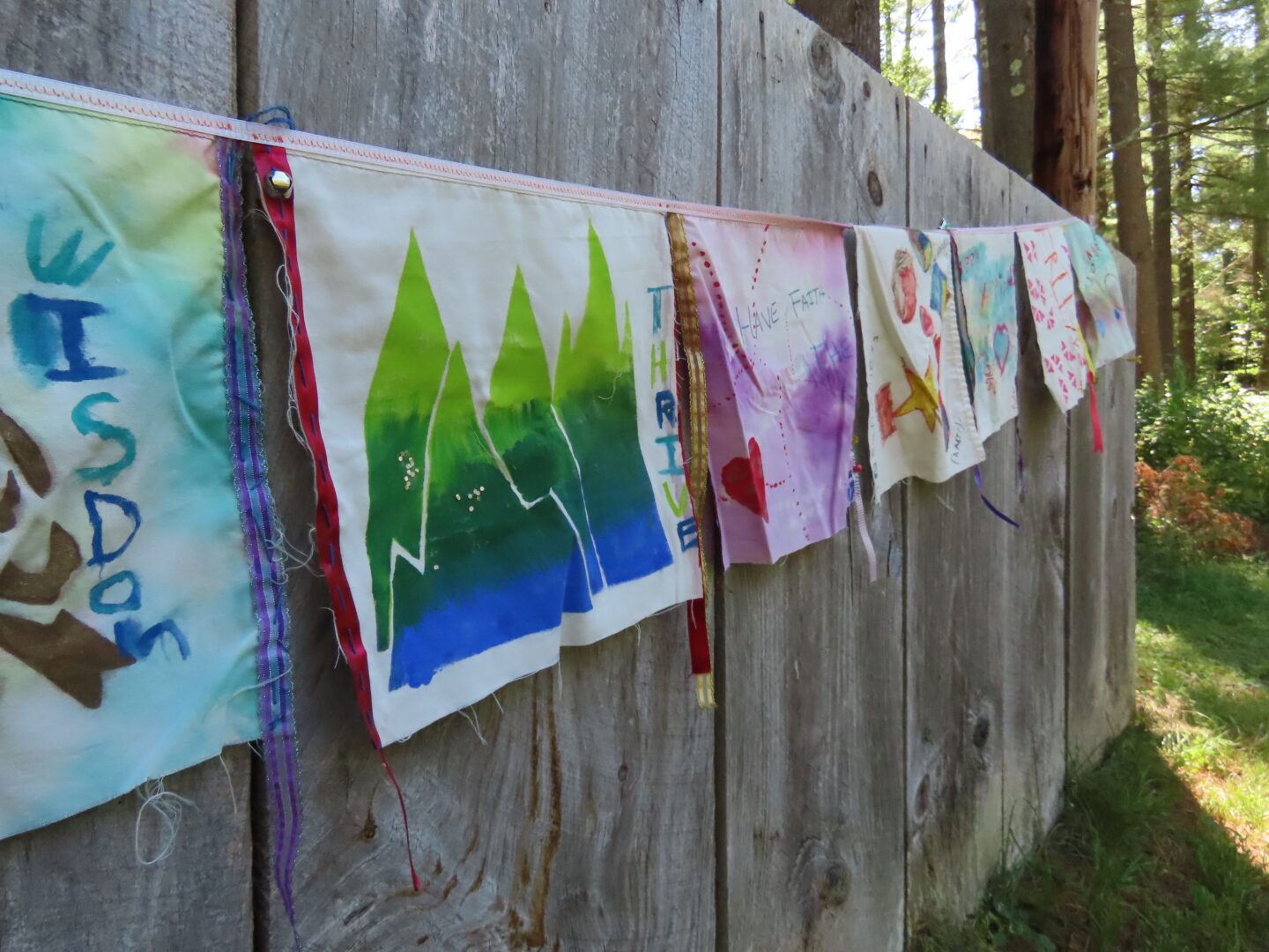 Squares of cloth painted with scenes such as a blue and green mountain scene hang on a string against a wooden wall.