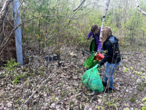 Two young women in jackets and jeans hold large green trash bags as they clean up a woodland area.