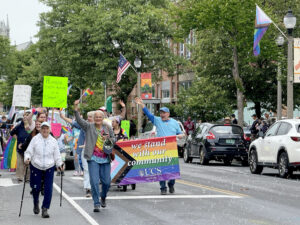 A large group of people marching down a city street. At the front of the group, a large Pride flag is carried by two people.