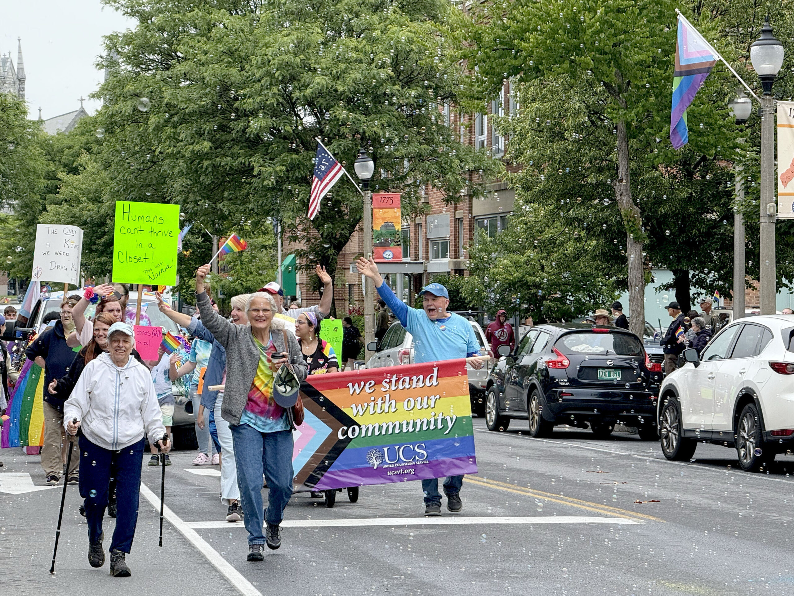 A large group of people marching down a city street. At the front of the group, a large Pride flag is carried by two people.